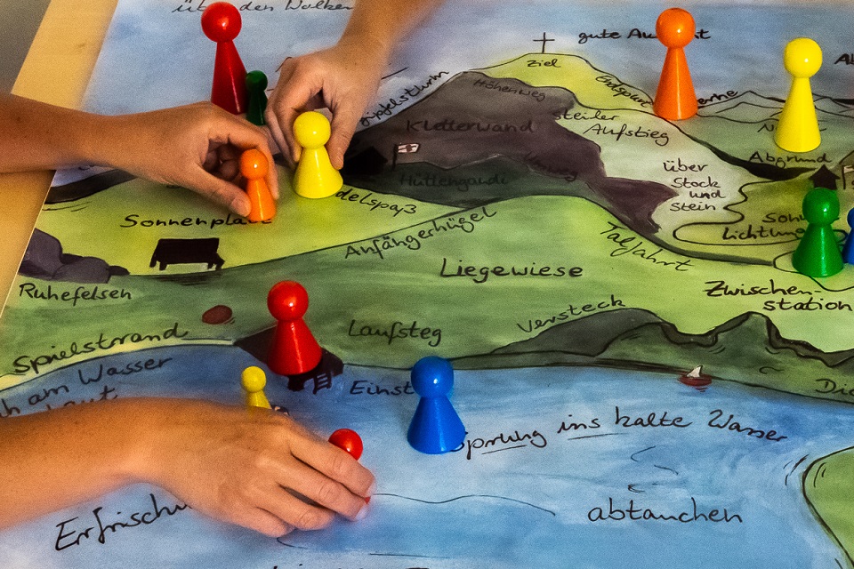 Das Bild zeigt bunte Spielfiguren auf einem Spielfeld mit aufgezeichneten Hügeln, Wiesen und Seen.