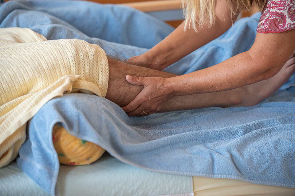 Das Bild zeigt die Beine eines Mannes eingebettet in Tücher. Die Hände einer Frau reiben die Beine mit Öl ein.