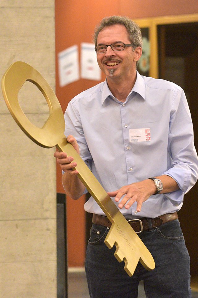 Das Bild zeigt einen Mann mit einem grossen goldenen Schlüssel aus Holz in der Hand.