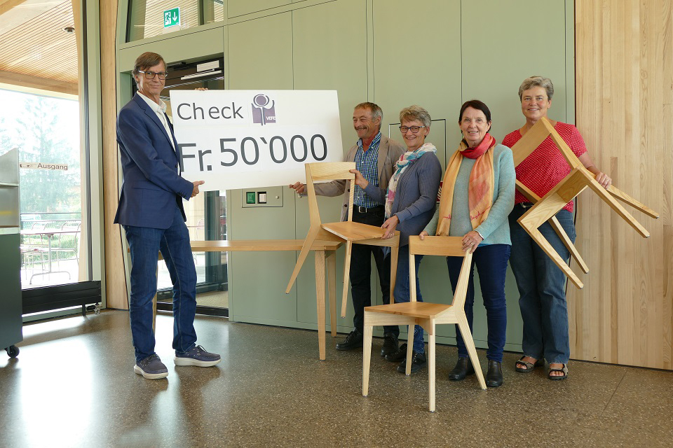 Das Bild zeigt zwei Männer die einen grossen Check mit der Aufschrift, 50 Tausend Franken, in die Kamera halten. Daneben stehen drei Frauen die moderne Holzstühle in den Händen halten.