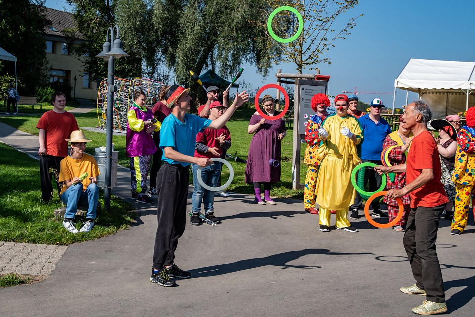 Das Bild zeigt zwei Männer die mit bunten Ringen jonglieren. Im Hintergrund stehen mehrere Clowns.
